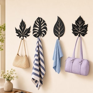 May include: Four black wall-mounted hooks shaped like various leaves. The hooks hold a beige woven handbag, a blue and white striped towel, a light blue cloth, and a lavender handbag. Each hook features a distinct botanical leaf silhouette.