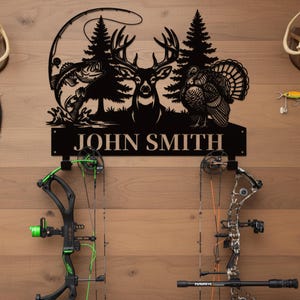 May include: Black metal wall art featuring a deer, turkey, and fish, with the text "JOHN SMITH". Two bows are mounted below the art. The bows are black and green, and black and brown.