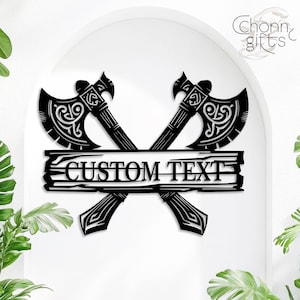 May include: Black metal wall art featuring two crossed axes with decorative handles and blades. A wooden plank with the words "CUSTOM TEXT" is centered below the axes. The axes have intricate designs, and the overall theme is rustic and personalized.