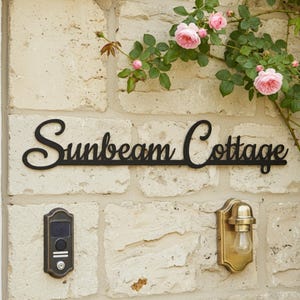 May include: A black metal sign reading "Sunbeam Cottage" in cursive lettering, fixed to a light stone wall. A brass light and doorbell are also mounted on the wall. Pink roses and green foliage are visible in the background.