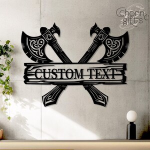 May include: Black metal wall art featuring two crossed axes with ornate detailing. A horizontal plank with the words "CUSTOM TEXT" is centered below the axes. The design is suitable for personalized home decor.