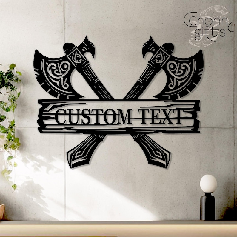 May include: Black metal wall art featuring two crossed axes with ornate detailing. A horizontal plank with the words "CUSTOM TEXT" is centered below the axes. The design is suitable for personalized home decor.