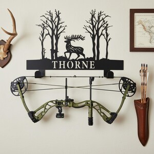 Può includere: Un espositore da parete in metallo nero per arco e frecce con il nome "THORNE" in lettere bianche. L'espositore presenta una silhouette di cervo e alberi. Un arco composto verde oliva è montato sotto, con una faretra in pelle che contiene frecce a destra.