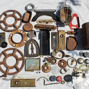 May include: A collection of vintage metal hardware and industrial components, including wheels, door handles, latches, and a light fixture. The items display a range of colors from rusty brown to tarnished brass, suggesting age and use. The objects are arranged on a white surface.