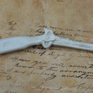 May include: A small, off-white replica of a flintlock pistol, resting on a vintage paper background with handwritten script and numbers. The miniature firearm is detailed, showcasing the trigger, hammer, and barrel.