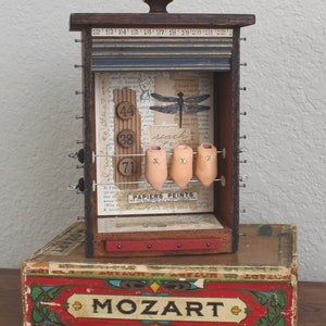 May include: A wooden box with a sliding door and a dragonfly design. The box has a number of small drawers and a row of small ceramic containers with the letters X, Y, and Z. The box is sitting on top of a red and gold box with the word "MOZART" printed on it.