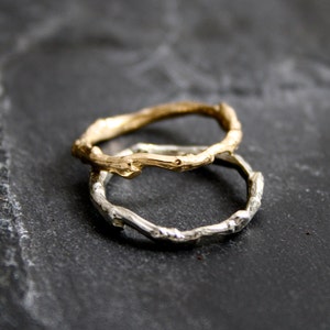 May include: Two twig-shaped rings, one gold and one silver, are stacked on top of each other. The rings are made to look like natural branches.