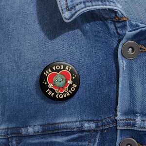 May include: A black button with a white border and a cartoon image of the Earth wearing a red heart. The text "See you at the equator" is written around the image.