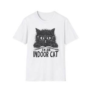 以下が含まれることがあります： 黒い眼鏡をかけ、本を読んでいる黒い猫が描かれた白いTシャツ。Tシャツには「私は室内猫です」と書かれています。