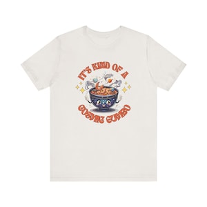 Könnte beinhalten: Weißes T-Shirt mit einer Cartoon-Illustration einer Schüssel mit kosmischem Gumbo. Die Schüssel hat ein lächelndes Gesicht und ist von Sternen und Planeten umgeben. Der Text "It's kind of a cosmic gumbo" ist in Orange und Rot gedruckt.
