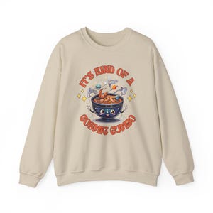Könnte beinhalten: Ein beiger Pullover mit einer Cartoon-Illustration einer Schüssel mit kosmischem Gumbo. Der Text "It's kind of a cosmic gumbo" ist auf dem Pullover aufgedruckt.