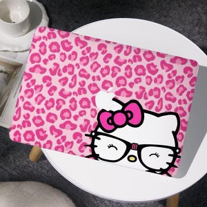 May include: A pink laptop case featuring a leopard print pattern. The case has a graphic of a white cat with a pink bow, glasses, and closed eyes. The laptop case is on a white table.
