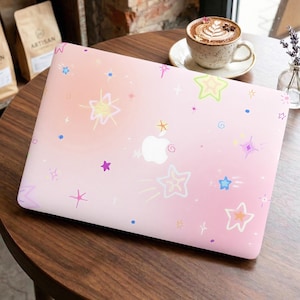 Può includere: Una cover per laptop rosa con effetto sfumato, ornata da illustrazioni colorate di stelle e brillantini. La cover presenta un logo Apple bianco ed è appoggiata su un tavolo di legno, accanto a una tazza di caffè e un piccolo vaso di fiori.