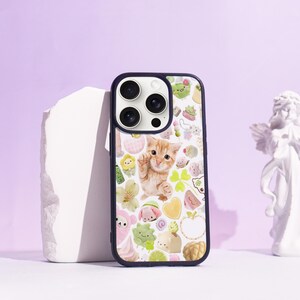 May include: A phone case with a navy blue border. The case features a light orange kitten surrounded by cartoon illustrations of flowers, hearts, and food items. The background is white, creating a whimsical design.