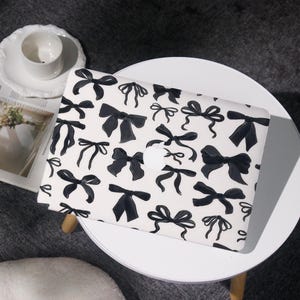 Può includere: Custodia bianca per laptop con un motivo di fiocchi neri. I fiocchi variano per dimensioni e stile, creando un design giocoso ma elegante. La custodia è su un tavolino bianco.
