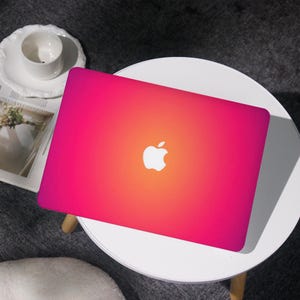 Könnte beinhalten: Ein Laptop mit einem rosa-orangenen Farbverlauf und dem Apple-Logo. Der Laptop steht auf einem kleinen weißen Tisch. Eine weiße Tasse und Untertasse sind im Hintergrund zu sehen.