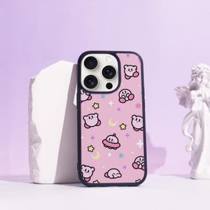 May include: A phone case with a pink background and a repeating pattern of cartoon characters, stars, and moons. The case has a black border and a camera cutout.