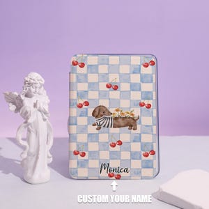 Peut inclure: Une coque de téléphone personnalisée avec un motif à carreaux bleu et blanc, représentant un teckel portant une chemise rayée et portant des fleurs. Des cerises rouges sont dispersées sur le design. Le nom "Monica" est imprimé sur la coque, avec le texte "CUSTOM YOUR NAME" en dessous.