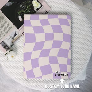 May include: A rectangular, lavender and cream-colored checkered pattern notebook cover. The text "Monica" is written in cursive, with the words "CUSTOM YOUR NAME" below it. The notebook is on a white, textured surface.