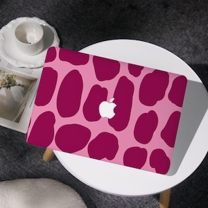 May include: A pink laptop skin featuring a pattern of large, irregular magenta shapes. The Apple logo is visible in white. The laptop is on a white table, with a cup and saucer and a magazine nearby.
