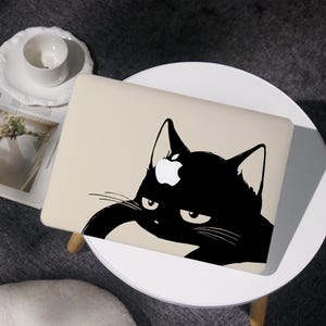 May include: A beige laptop with a black cat graphic. The cat has a white Apple logo on its head and a grumpy expression. The laptop is on a white table with a cup and saucer.