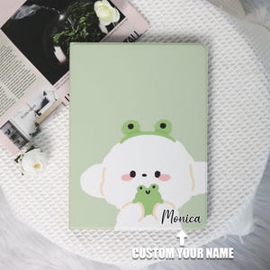 Op de afbeelding: Een groene en witte geïllustreerde hoes met een schattig cartoonhondje dat een kikker vasthoudt. De hoes heeft de naam "Monica" erop en de tekst "CUSTOM YOUR NAME" eronder.