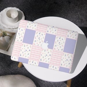 Peut inclure: Un ordinateur portable avec un motif patchwork rose pastel, lavande et blanc. Le motif comprend des imprimés floraux, des pois et un motif à carreaux. Le logo Apple est visible.