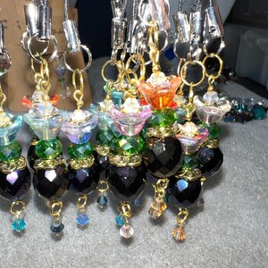 May include: A collection of colorful beaded charms with black crystal beads and gold accents. The charms feature flower-shaped beads in shades of pink, blue, green, and orange. The charms are attached to silver and gray cords.