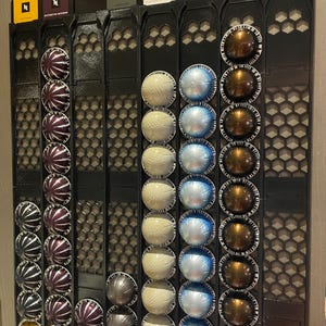 Può includere: Un organizzatore da parete in metallo nero con cinque file di capsule di caffè. Le capsule sono disposte in file per colore: oro, viola, bianco, blu e marrone.