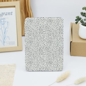 May include: A rectangular tablet case with a white background and a black speckled pattern. The case is standing upright on a white surface. Other items in the background include a framed picture, a small plant in a white pot, and a cardboard box.