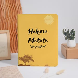 May include: A rectangular yellow notebook featuring a stylized sun graphic in the top right corner, a brown silhouette of a tree at the bottom, and the black text "Hakuna Matata" followed by the English translation "No problem" in the center.
