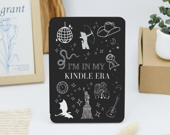 May include: A black e-reader cover featuring white line art illustrations of a disco ball, archer, cowboy hat, snake, ribbon, clock, Statue of Liberty, couple, guitar, and dragon, with the text "I'M IN MY KINDLE ERA" printed in the center.