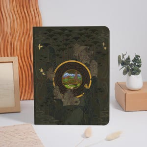 May include: A hardback journal with a dark green cover featuring an illustration of a landscape scene within a gold circular frame. The cover also includes illustrations of figures, trees, and birds. The journal is displayed on a white surface.