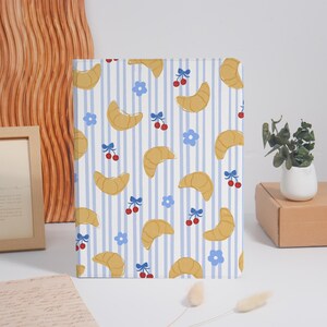 May include: A rectangular tablet case featuring a repeating pattern of golden-brown croissants, small red cherries with blue stems, and light blue flowers set against a background of vertical white and light blue stripes.