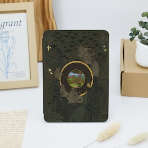 May include: A dark green Kindle cover featuring a gold and green illustration of a tree and a path leading to a mountain range. The illustration is encircled by a gold circle with a black and gold pattern.