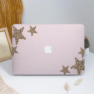 May include: A light pink laptop case with a white Apple logo and several star-shaped leopard print decals. The case is on a white surface with a wooden frame and decorative elements in the background.