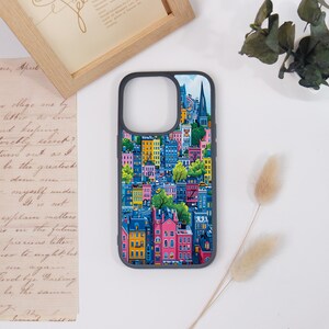 May include: A phone case with a colorful illustration of a cityscape. The case has a dark gray border and features buildings in shades of pink, yellow, blue, and green. The design includes trees and a church steeple.
