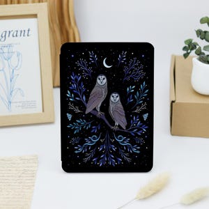 Op de afbeelding: Een zwarte e-reader hoes met een gedetailleerde illustratie van twee uilen op een tak. Het ontwerp bevat blauw en paars gebladerte, kleine witte sterren en een maansikkel tegen een donkere achtergrond, wat een nachtelijk tafereel creëert.