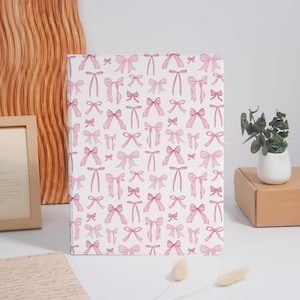 May include: A white notebook with a repeating pattern of pink bows. The bows vary in style and size, creating a delicate and feminine design. The notebook is rectangular and appears to be closed.