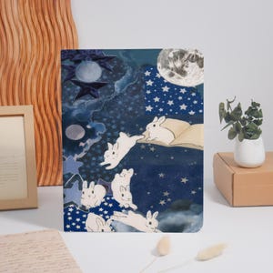 May include: A decorative notebook with a whimsical night sky design. The cover features white rabbits, stars, and a full moon against a dark blue background. The notebook is a unique piece of stationery.