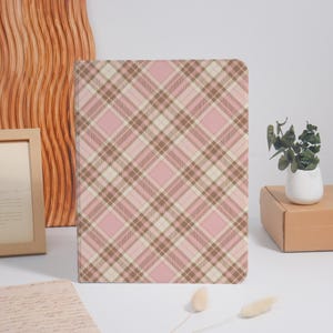 May include: A pink, brown, and cream plaid pattern notebook. The notebook has a soft pink background with intersecting lines in brown and cream, creating a classic plaid design. The notebook is rectangular and appears to be closed.