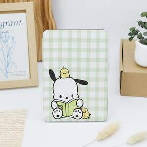 May include: A rectangular e-reader case featuring a light green and white gingham pattern. The front displays a white cartoon dog character with black ears reading a green book, with a small yellow chick on its head and another beside it.