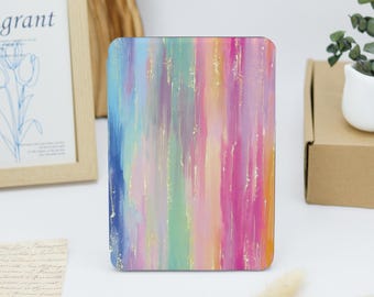 May include: A rectangular, colorful case with a vertical brushstroke design in shades of blue, green, pink, and gold. The case is standing upright on a white surface. The case has rounded corners.