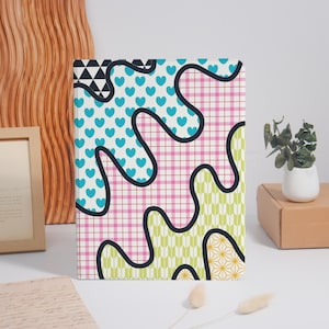 May include: A patterned notebook with a striking design. The cover displays a mix of patterns, including blue hearts, pink gingham, and geometric shapes, all outlined in black. A vibrant accessory.