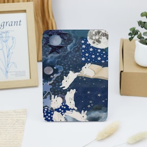 Peut inclure: Un collage bleu et blanc représentant une scène fantaisiste de lapins blancs volant dans un ciel nocturne étoilé. Les lapins volent au-dessus d'un livre avec une lune et des étoiles en arrière-plan.