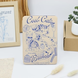 May include: A beige Kindle cover with a blue and white illustration of cats enjoying brunch. The text reads "Cool Cats are always getting Brunch! Can't keep those paws off the mimosas."