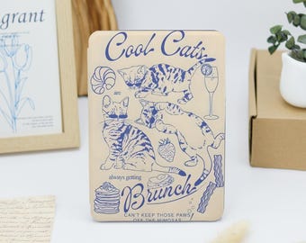 May include: A beige Kindle cover with a blue and white illustration of cats enjoying brunch. The text reads "Cool Cats are always getting Brunch! Can't keep those paws off the mimosas."