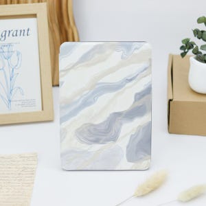May include: A rectangular e-reader case featuring a marbled pattern with flowing diagonal bands of white, light gray, and soft beige. The surface has a smooth finish and covers the front of the device, displaying a fluid, abstract design throughout.