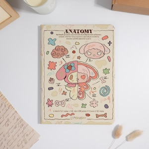 K&ouml;nnte beinhalten: Ein Notizbuch mit beigem Einband, das eine Cartoon-Illustration eines Charakters zeigt, der in zwei H&auml;lften geteilt ist und die Anatomie zeigt. Der Einband enth&auml;lt das Wort "ANATOMY" und verschiedene bunte Kritzeleien und Etiketten. Das Notizbuch liegt auf einer wei&szlig;en Oberfl&auml;che.