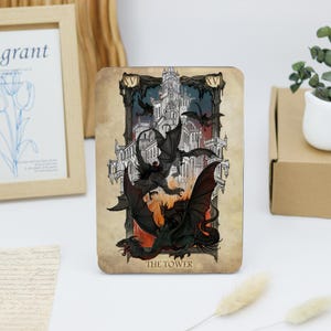 May include: A tarot card with a dark background and a white tower in the center. The tower is surrounded by four black dragons with red flames at the base. The card is labeled "The Tower" and has the number XV at the top.